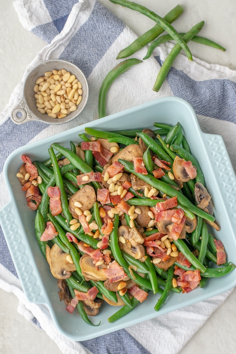Sauteed green beans with mushrooms and bacon are topped with pine nuts